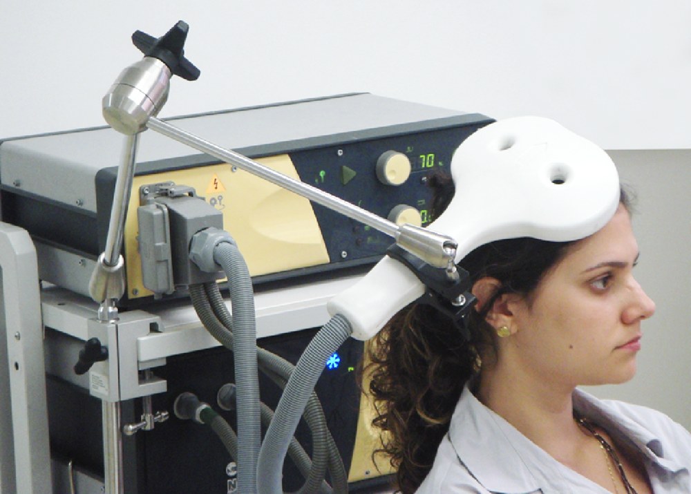 A person seated beside a Neuro-MS TMS device, with a coil near the head